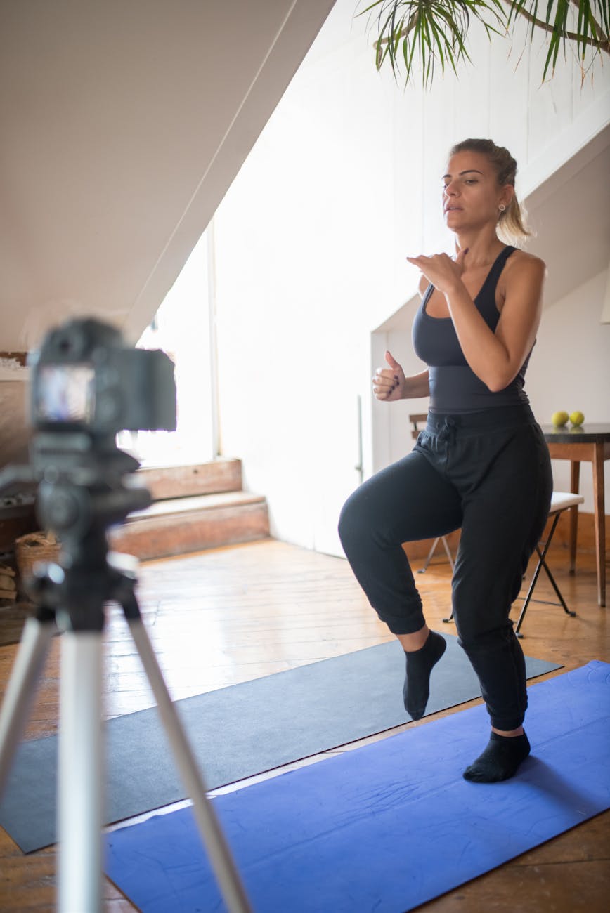 A woman demonstrating an exercise on a blue workout mat in a home setting, with a camera positioned on a tripod capturing the session.